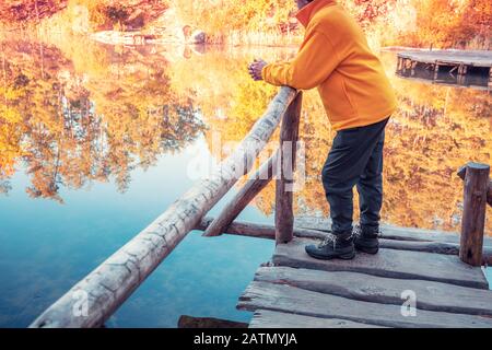 Un uomo si erge su un ponte di legno appoggiato su una ringhiera e guarda ad un lago forestale. Alba sul lago in autunno Foto Stock