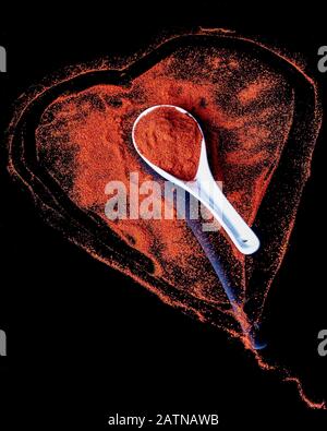 Bella composizione con un cucchiaio bianco di ceramica pieno di rosso forte paprika con una forma cuore rosso su una base nera lucida sullo sfondo Foto Stock