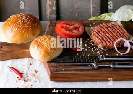 Ingredienti per la preparazione di hamburger fatti in casa su tagliere di legno, serviti con forchetta di carne e coltello sopra tovaglia bianca. Stile rustico scuro. Foto Stock