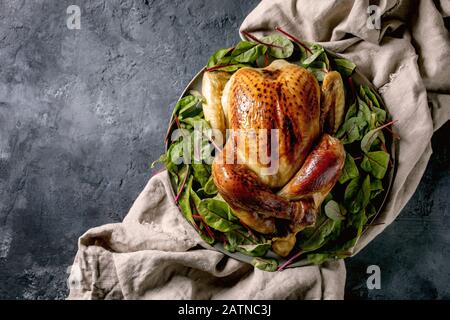 Pollo intero cotto su piatto di ceramica, servito con foglie di insalata verde di barbabietole su tela di lino su fondo di cemento nero. Disposizione piatta, spazio. Foto Stock