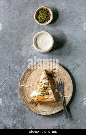 Pezzo di cheesecake con condimento del latte e pistacchi su piastra in ceramica con forchetta e salsa in recipiente in ceramica su sfondo grigio texture. Piatto, spac Foto Stock