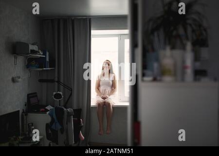 Giovane donna pensiva seduto alla finestra che guarda il suo riflesso nel grande specchio a casa interni moderni, rilassante e sognante Foto Stock