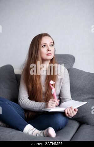 Giovane femmina creativa e penosa seduta sul divano a casa scrivere qualcosa nel suo diario con divertente penna rosa in forma di fenicottero. Donna attraente Foto Stock