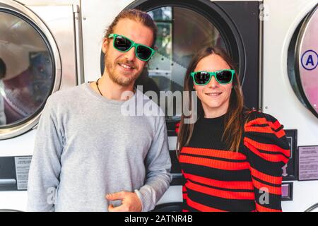 giovane coppia loving che indossa occhiali da sole verdi sta aspettando i vestiti di lavaggio in una lavanderia pubblica Foto Stock