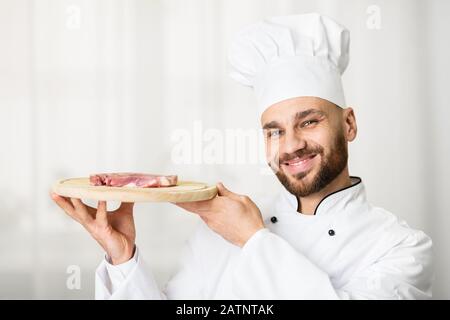 Maschio Chef Holding Bistecca Fresca Su Tagliere A Tavola In Cucina Foto Stock