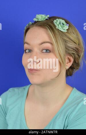 ritratto di una giovane bionda scettica donna caucasica con capelli opacici e fiori cian su un pelo circlet ruminante di fronte a sfondo blu Foto Stock