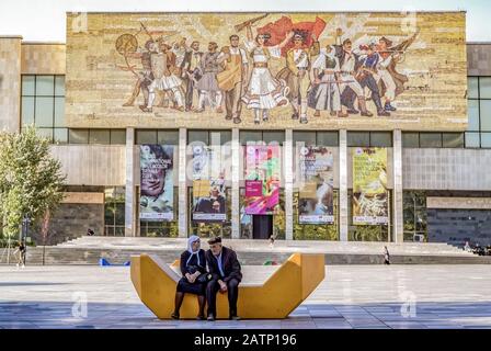 Coppia albanese anziana seduta sulla piazza Skanderbeg di fronte al Museo Nazionale di Storia. Mosaico che mostra gli albanesi vittoriosi della storia. Foto Stock