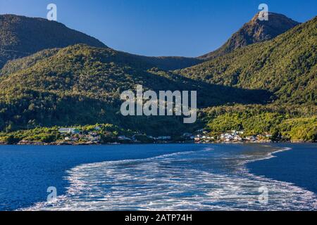 Scia del traghetto Quelat che lascia il villaggio di Puerto Gaviota a Isla Magdalena, montagne coperte di foresta pluviale temperata, regione di Aysen, Patagonia, Cile Foto Stock