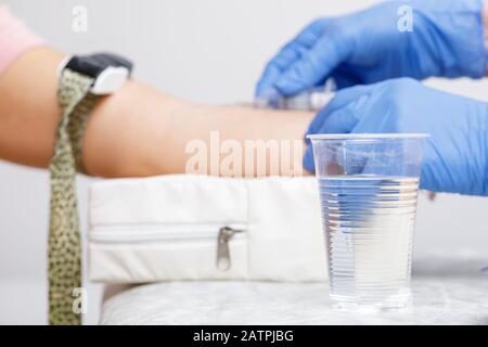 Paura della procedura di prelievo dei campioni di sangue concetto con il medico che raccoglie il sangue dalla vena del braccio di un paziente in background e tazza di acqua in plastica a fuoco Foto Stock