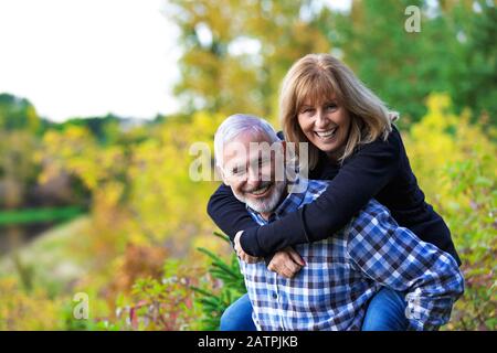 Un huband maturo che si diverte e che dà a sua moglie un cavalcare indietro su un sentiero lungo un fiume in un parco cittadino in una calda serata autunnale Foto Stock