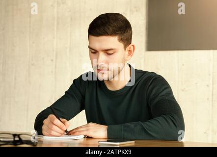 Giovane Uomo Che Prende Appunti Che Lavora Seduto In Ufficio Foto Stock