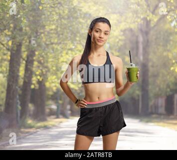 Giovane donna in abbigliamento sportivo che tiene un frullato verde e posa in un parco Foto Stock