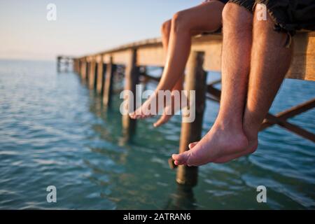 Due coppie di gambe penzolanti off di un molo in legno. Foto Stock