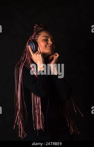 Bella ragazza con pigtail africani zizi ascolta con occhi chiusi la musica in cuffie su uno sfondo nero. Emozioni di felicità, piacere e p Foto Stock