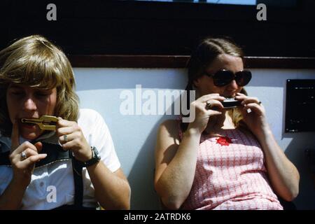 Fotografia vernacolare scattata su un film analogico 35mm trasparenza, si ritiene di raffigurare la donna in bianco t-shirt e occhiali da sole nero seduta su sedia, con l'uomo accanto a lei, entrambi giocando armonicas, 1970. Gli argomenti/oggetti principali rilevati includono l'azione umana e la musica. () Foto Stock