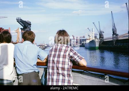 Fotografia vernacolare scattata su un film analogico 35mm trasparenza, si ritiene di raffigurare l'uomo in rosso e bianco camicia vestito plaid seduta su barca di legno marrone durante il giorno guardando verso gru di un porto commerciale, 1970. I principali argomenti/oggetti rilevati includono Barca, Veicolo, Moto D'Acqua e mare. () Foto Stock