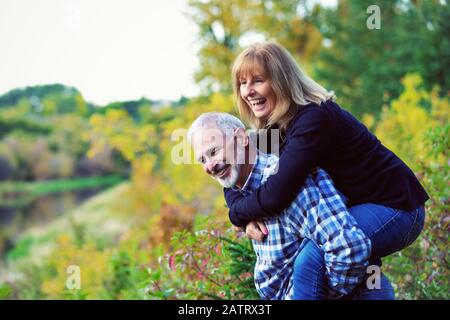 Un huband maturo che si diverte e che dà a sua moglie un cavalcare indietro su un sentiero lungo un fiume in un parco cittadino in una calda serata autunnale Foto Stock