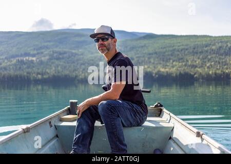 L'uomo si siede in una piccola barca a remi con motore sul Lago Bianco con l'acqua tranquilla che riflette la foresta sullo sfondo; British Columbia, Canada Foto Stock