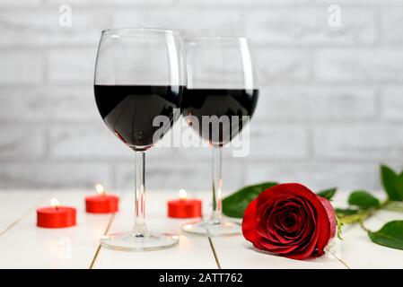 Concetto di San Valentino. Due bicchieri di vino, rosa rossa e candele accese su un tavolo di legno bianco. Foto Stock