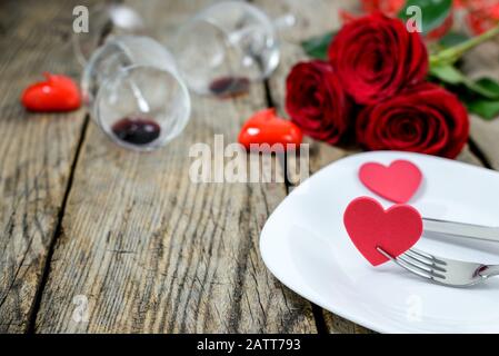 Cuori su piastra con forchetta e coltello, due bicchieri di vino, rose rosse e candele su un vecchio tavolo di legno. Messa a fuoco selettiva. Foto Stock