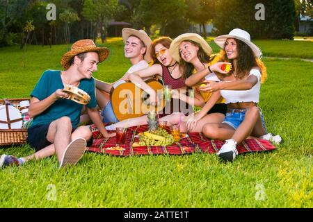 Felici i giovani amici che hanno picnic estate nel parco, seduti insieme su tappeto rosso in cappelli di paglia, cantare e suonare su chitarra e tamburello. Foto Stock