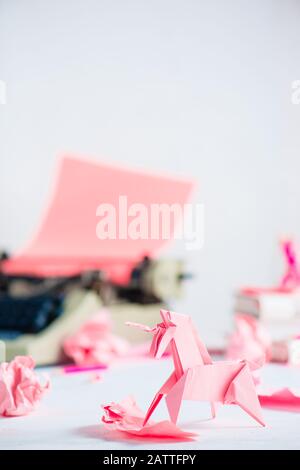 Unicorno di origami rosa con palline di carta sbriciolate e una macchina da scrivere. Processo creativo per iscritto Foto Stock