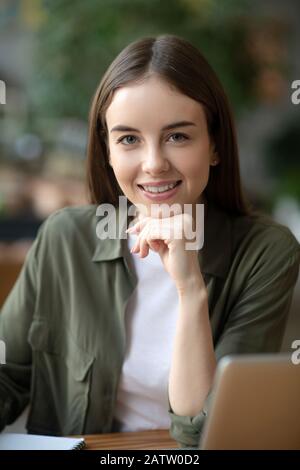 Emozione positiva. Bella ragazza di fronte a un computer portatile che guarda di fronte a se stessa e sorridente, propping il mento con la mano. Foto Stock