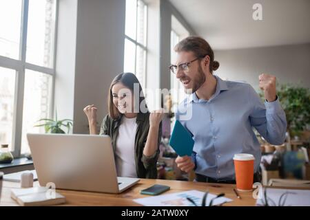 Questa è la vittoria. Giovane entusiasta ragazza e uomo in occhiali e con un tablet in mano, in piedi di fronte a un laptop e guardando lo schermo, braccia r Foto Stock