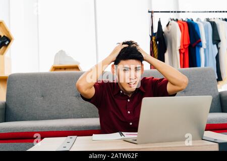 Scioccato giovane asiatico in abbigliamento casual con bocca aperta guardando rapporto di progetto su laptop seduto al tavolo a casa Foto Stock