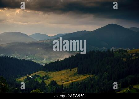 Tramonto d'oro sulle montagne dei carpazi - bellissimo paesaggio estivo, spiuces su colline, villaggio, case, cielo nuvoloso scuro e luce solare brillante, prato e Foto Stock