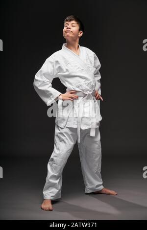 un adolescente vestito in abiti di arti marziali pone su uno sfondo grigio scuro, un concetto sportivo Foto Stock