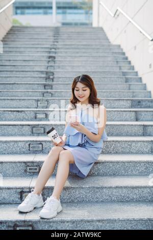 Bella giovane donna asiatica seduta al gradino esterno e utilizza lo smartphone. Lo stile di vita della donna moderna. Foto Stock