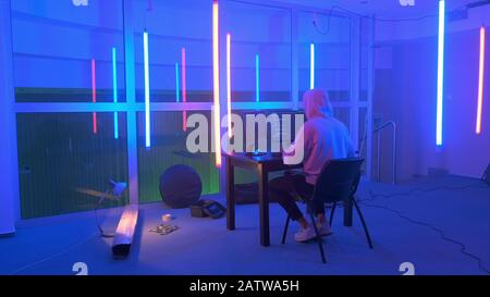 Hacker in bianco hoodie penetrante sistema di rete in sala neon. Intorno al tavolo c'è un'attrezzatura diversa. Il pirata informatico maschile in una cappa funziona su un computer con dati sugli schermi del display. Foto Stock