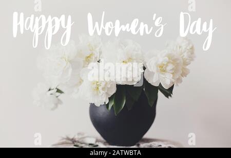 Happy Women's day text scritto a mano su elegante bouquet di peonia in vaso di argilla nera su tessuto di lino con forbici su sfondo rustico. Peonie bianche rurale Foto Stock