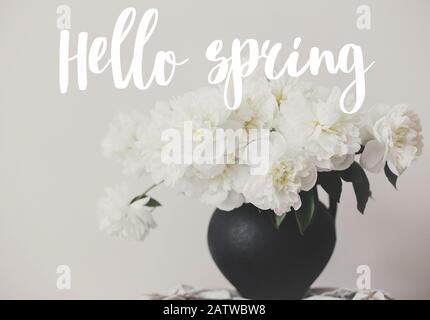 Ciao testo primavera scritto a mano su elegante bouquet di peonia in vaso di argilla nera su sfondo rustico. Peonie bianche rurale ancora vita. Primavera Foto Stock