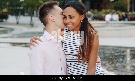 Primo piano di giovani che sussurrano qualcosa nell'orecchio della ragazza seduto su una panchina all'aperto. Sorridono, abbracciano e parlano l'uno con l'altro. Foto Stock