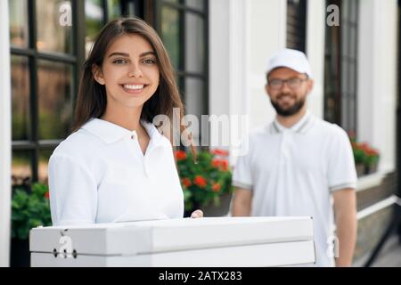 Vista frontale di felice giovane femmina gioiosa in blusa bianca che porta pizza e ragazzo in piedi dietro. Bella donna bruna che tiene due scatole pizza, guardando la macchina fotografica, ridendo. Concetto di consegna di cibo. Foto Stock