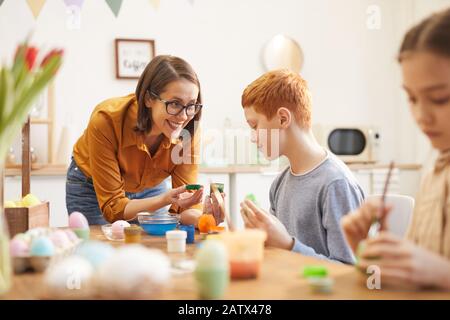 Sorridente madre che apre vernice verde e aiuta i suoi bambini a dipingere le uova per Pasqua al tavolo Foto Stock