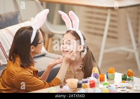 Madre giovane che dipinge il volto di sua figlia che fanno il costume di coniglio per la festa di Pasqua a casa Foto Stock