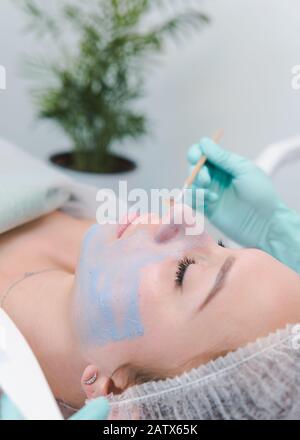 Il cosmetologo femminile che applica la maschera di cosmetologia blu al volto della giovane donna, primo piano. Trattamento viso cosmetico in clinica di bellezza. Concetto di pelle e. Foto Stock