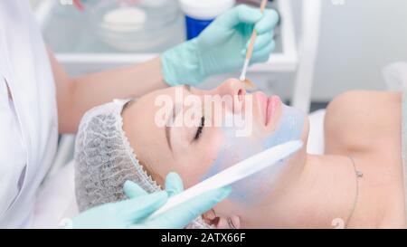 Il cosmetologo femminile che applica la maschera di cosmetologia blu al volto della giovane donna, primo piano. Trattamento viso cosmetico in clinica di bellezza. Concetto di pelle e. Foto Stock