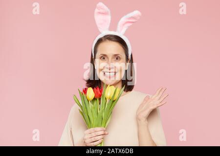 Ritratto di giovane donna felice in orecchie di coniglio che tiene bouquet di tulipani e sorridente alla macchina fotografica contro lo sfondo rosa Foto Stock