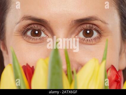 Primo piano del volto femminile con occhi marroni che guardano la fotocamera dietro i fiori Foto Stock