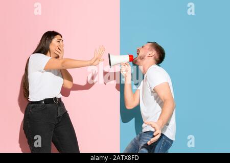 Chiama con il megafono, è arrabbiata. Giovane e felice uomo e donna in abiti casual su sfondo rosa, blu bicolored. Concetto di emozioni umane, expation facciale, relazioni, annuncio. Bella coppia. Foto Stock