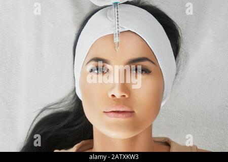 Vista dall'alto dell'iniezione di botox in testa di forehead femminile. Cosmetologo che usa una siringa con liquido speciale mentre bruna il paziente in un asciugamano sulla testa sdraiata, guardando la fotocamera. Concetto di cosmetologia, bellezza. Foto Stock