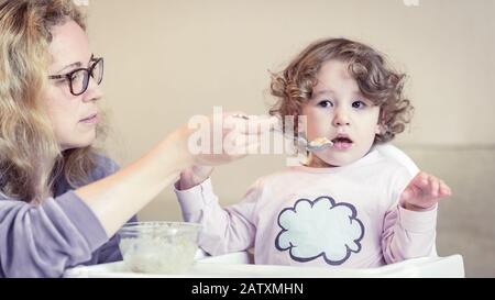 Bambina durante il consumo a casa. Madre nutre il suo bambino carino con un cucchiaio. Adorabile bambino siede su sedia alta e ha problemi con il cibo. I due anni- Foto Stock