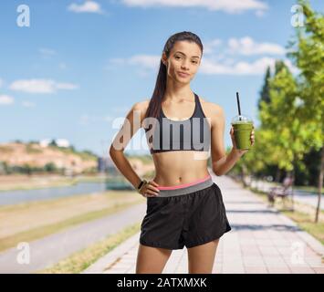 Giovane donna in abbigliamento sportivo che tiene un frullato verde su una pista da jogging vicino a un fiume Foto Stock