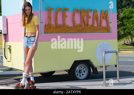 Bella ragazza teenage si trova sul pavimento indossando giallo brillante moderno top e vecchi pattini a rulli retro con lacci rossi accanto caravan retro con gelato Foto Stock