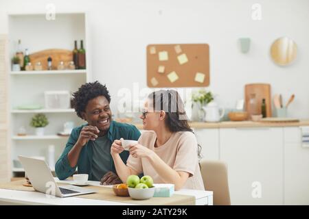 Giovane coppia felice seduto al tavolo di fronte al laptop bere caffè e sorridere l'uno all'altro in cucina Foto Stock