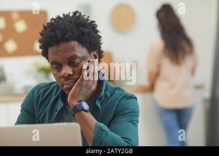 L'uomo africano serio che si concentra sul lavoro sul laptop mentre la donna cucina in background Foto Stock